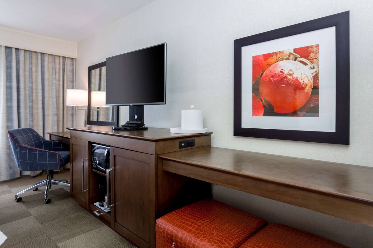 A hotel room with a wooden desk, a flat-screen TV, a chair, a lamp, a framed picture, and a minibar.