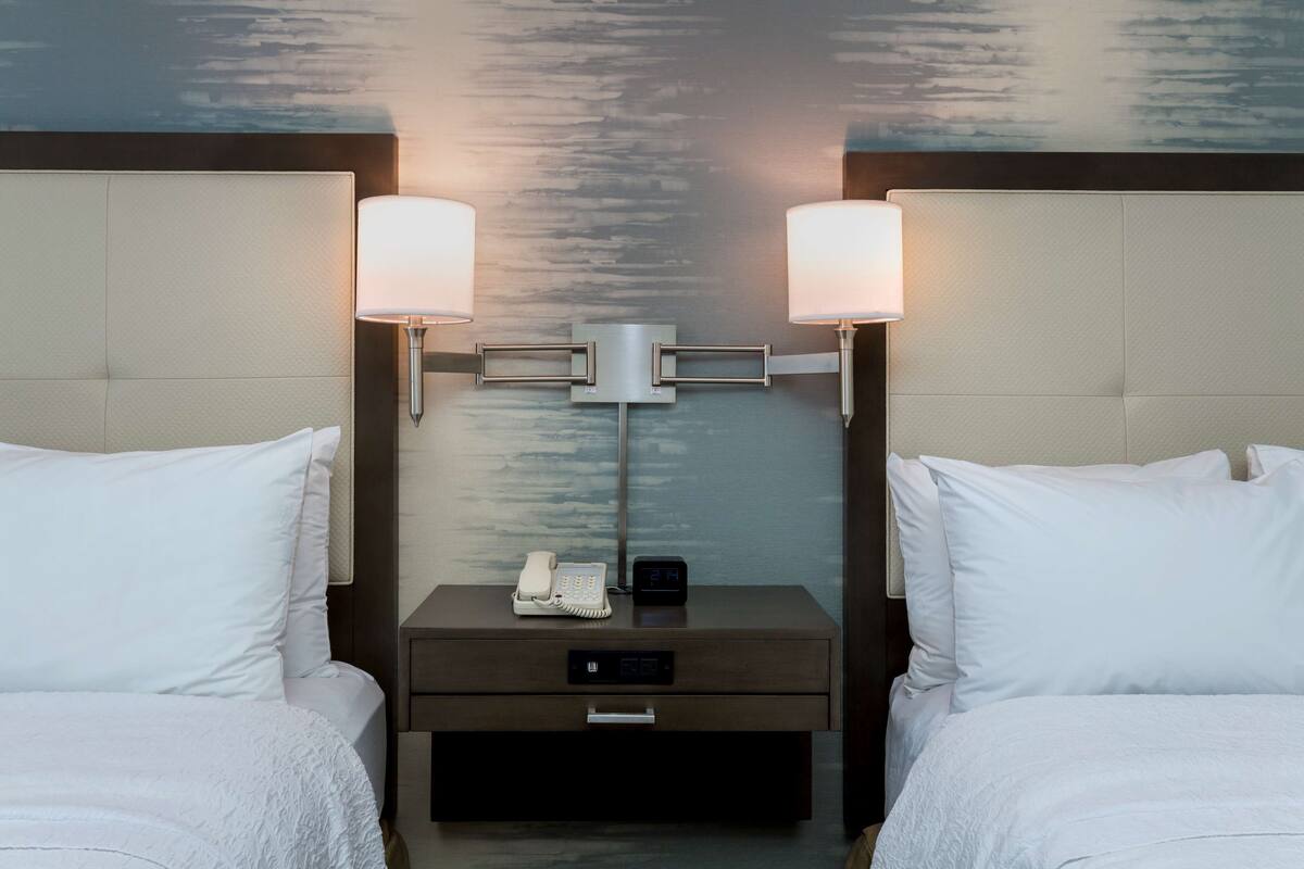 A hotel room with two beds, a nightstand with a lamp, a phone, and a digital clock.