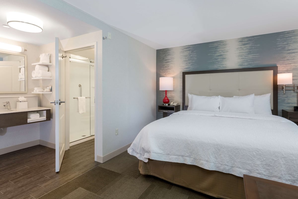 A hotel room with a large bed, a bathroom with a shower, and a bedside table with a lamp.