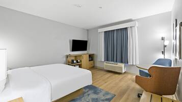 A hotel room with a bed, a desk, a chair, a TV, a window with curtains, and a heater.