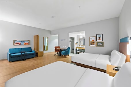 A modern hotel room with a large bed, a blue sofa, a wooden side table, and a desk with chairs.