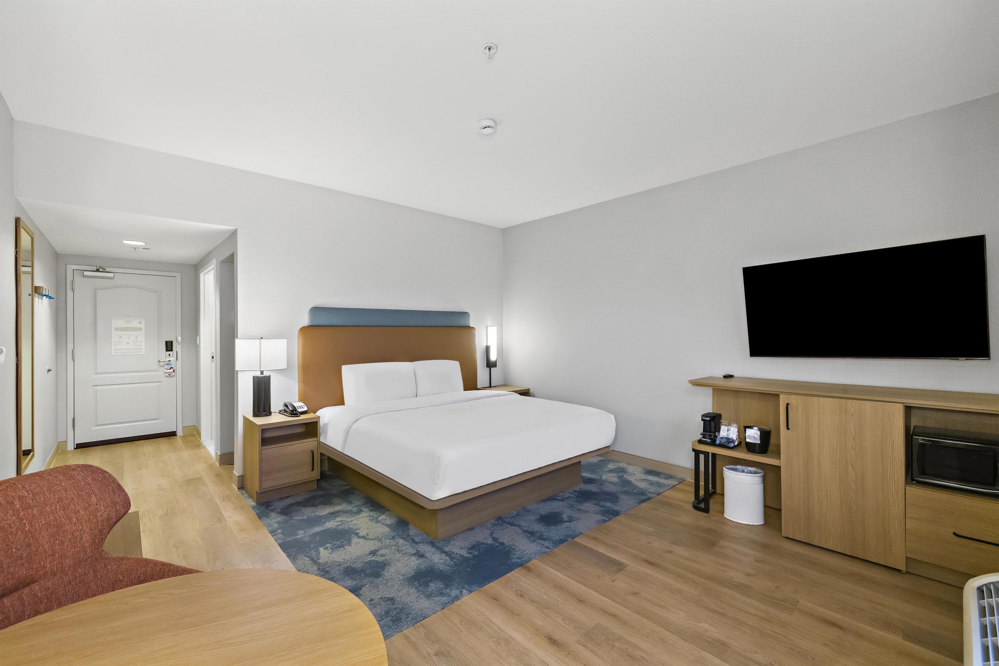A hotel room with a bed, bedside table, lamp, TV, and a small desk with a microwave.