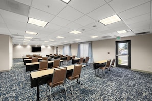 A conference room with rows of tables and chairs, a flat-screen TV on the wall, and a door leading outside.