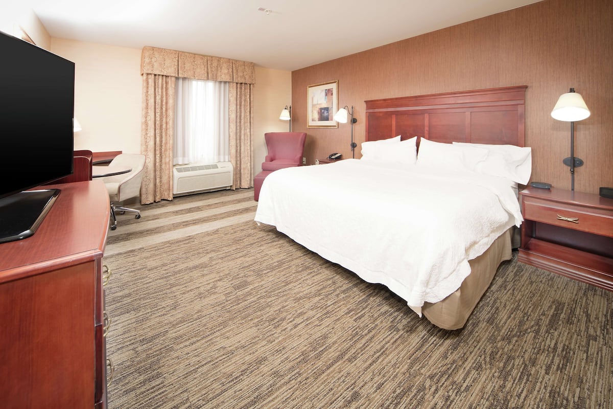 A hotel room with a large bed, a desk with a TV, a chair, a desk lamp, and a window with curtains.