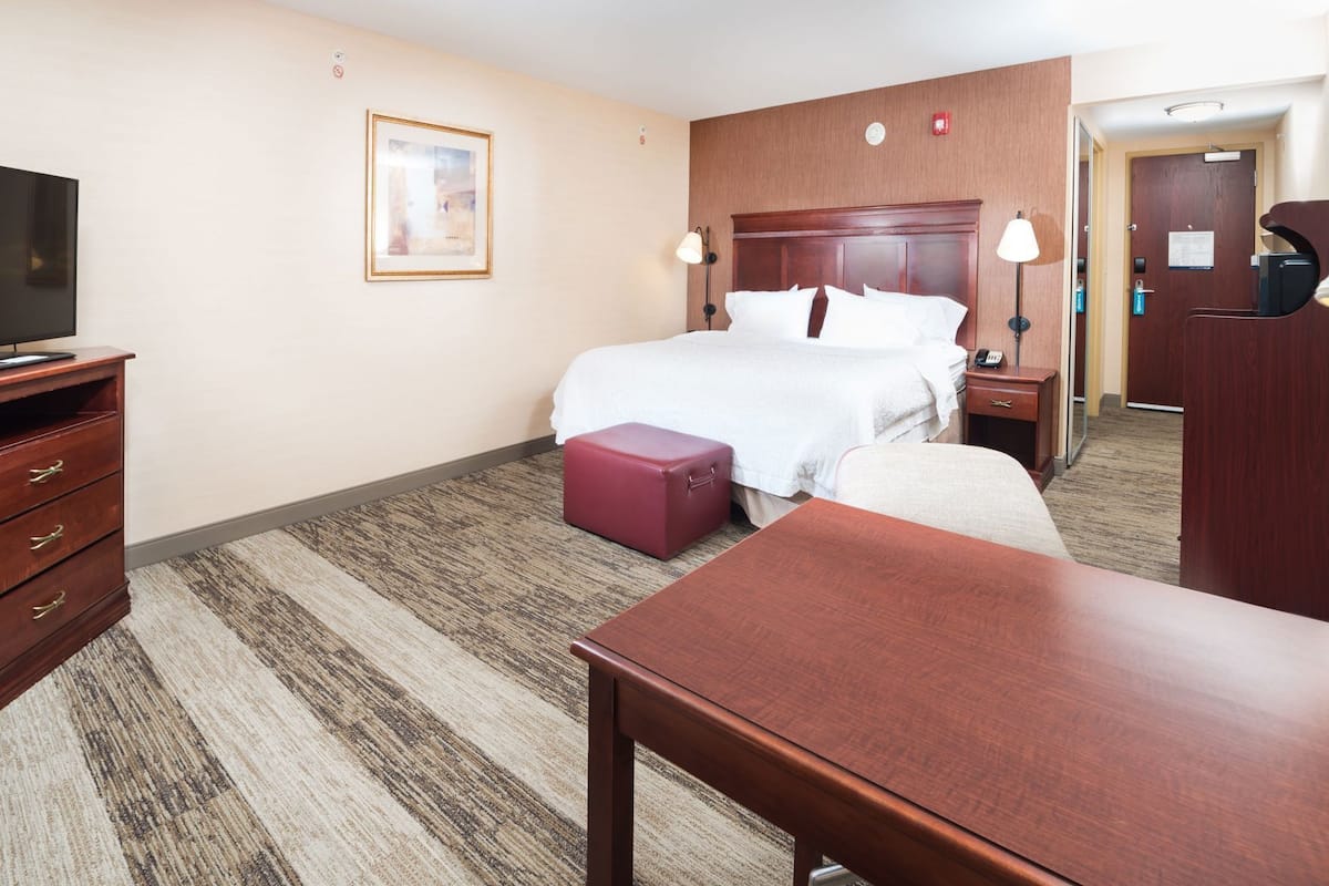 A hotel room with a bed, a desk, a dresser, a red suitcase, a framed picture on the wall, and a door leading to the bathroom.