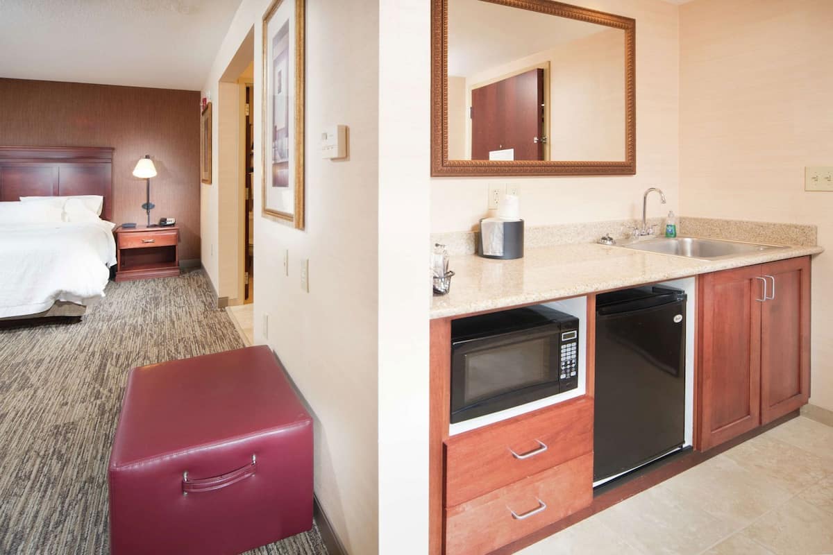 A hotel room with a bed, nightstand, lamp, and a red suitcase. The adjacent room has a kitchen with a microwave, dishwasher, sink, and cabinets.
