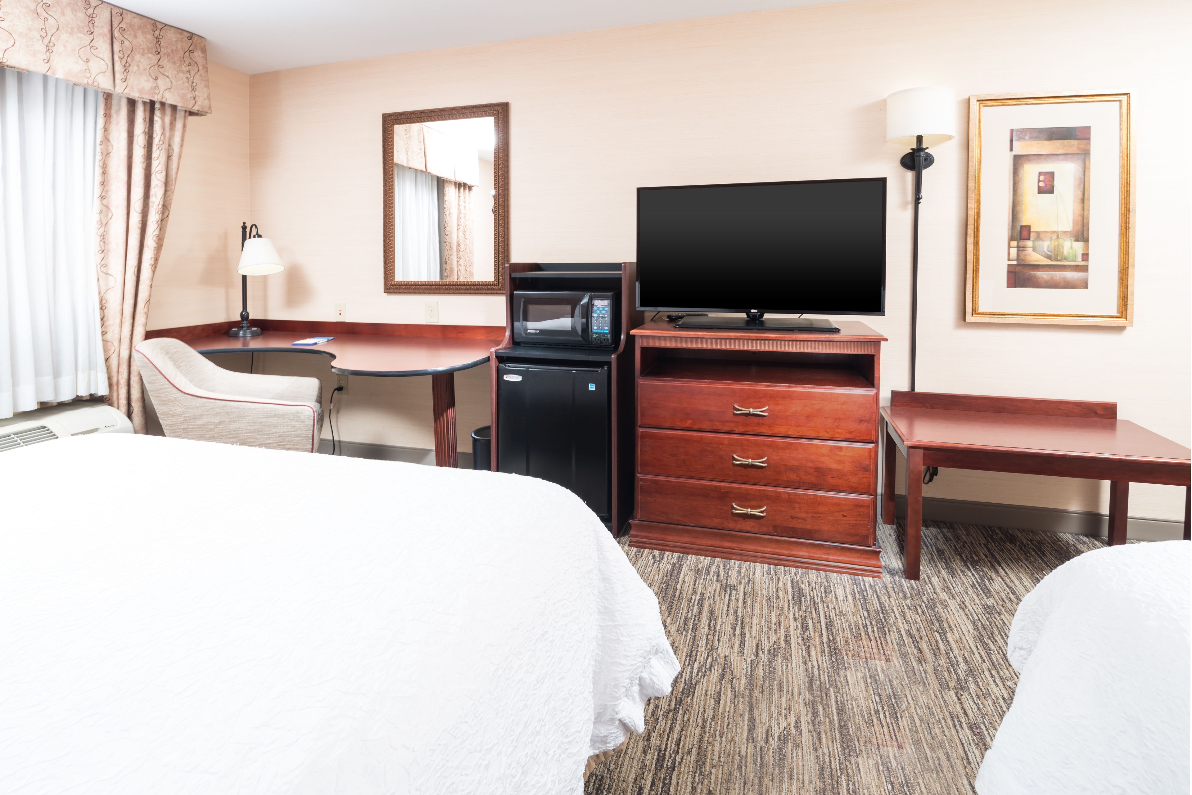 A hotel room with two beds, a desk, a television, a microwave, a dresser, and a lamp.