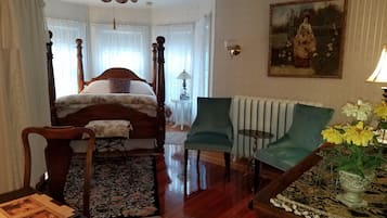 A bedroom with a four-poster bed, a green armchair, a small table with a vase of flowers, and a painting on the wall.
