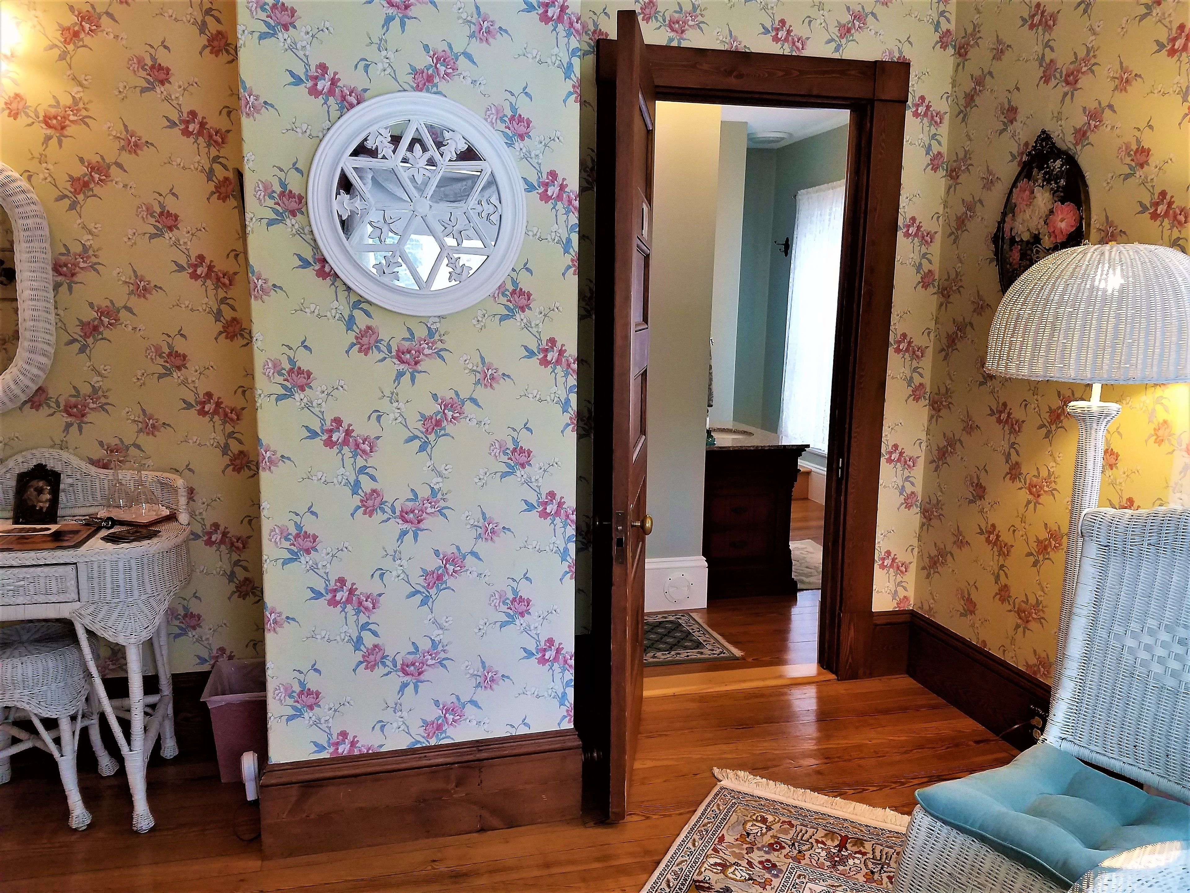 A room with floral wallpaper, a round mirror, wicker furniture, and a lamp.