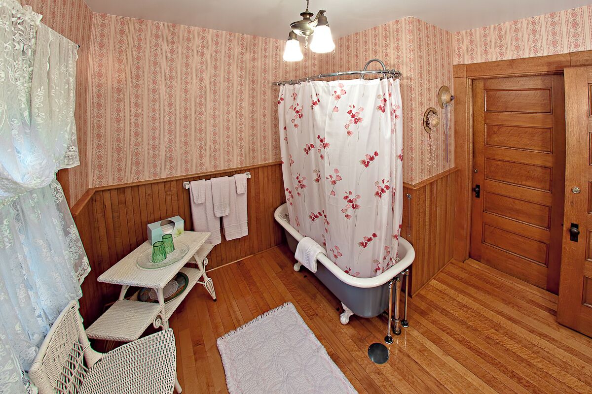 Une salle de bain avec un plancher en bois, une baignoire blanche agrémentée d’un rideau de douche à motifs floraux, un lavabo blanc et une porte en bois.