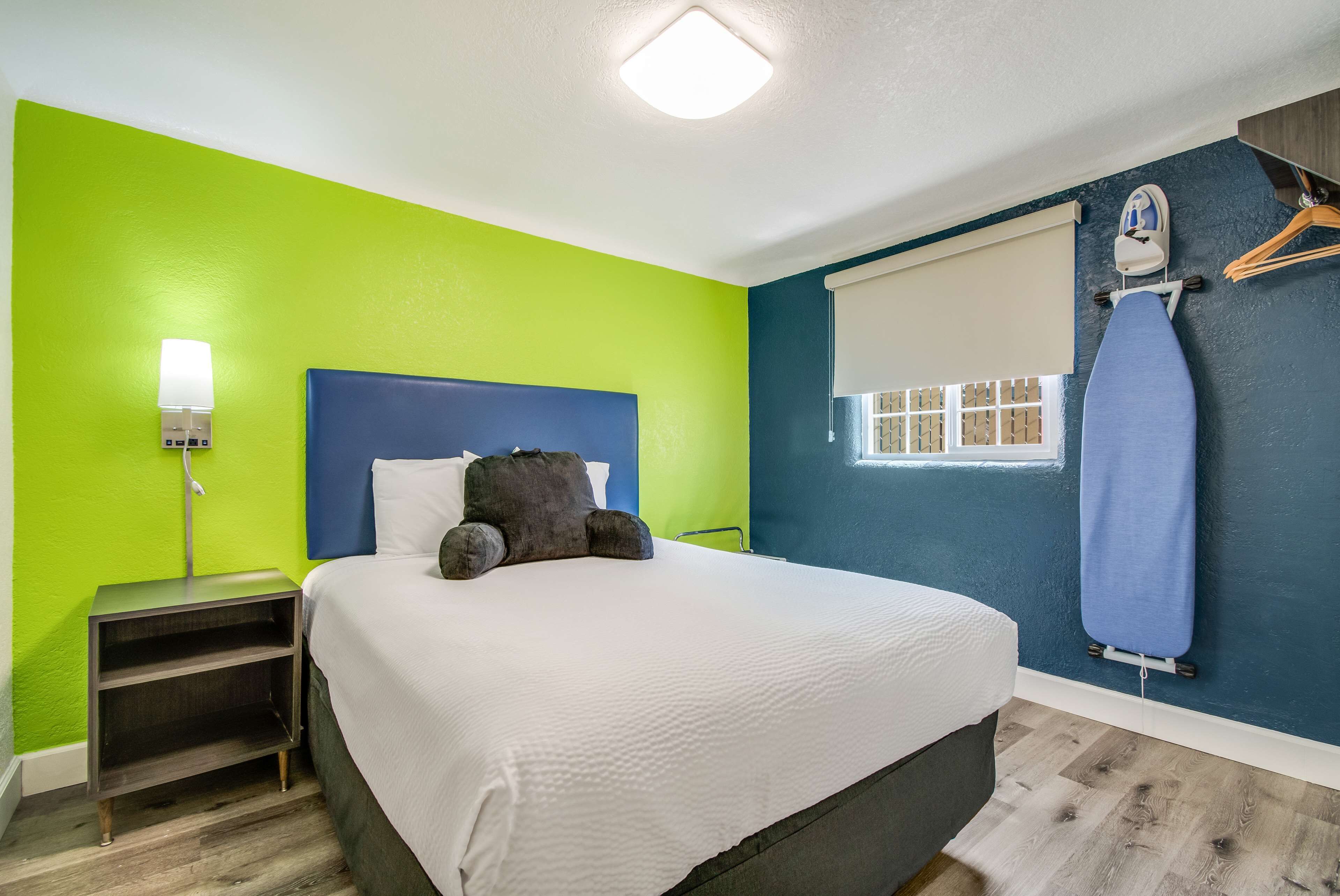 Una cama bien hecha con edredón blanco y cabecero oscuro en una habitación con paredes verdes y azules, una mesita de noche con lámpara y una ventana con vista a un muro de ladrillo.