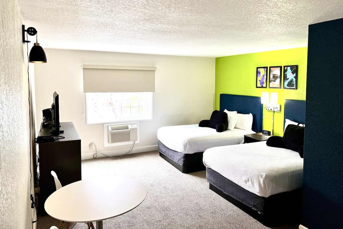 A hotel room with two beds, a desk, a chair, a table, a window with a view, and artwork on the wall.