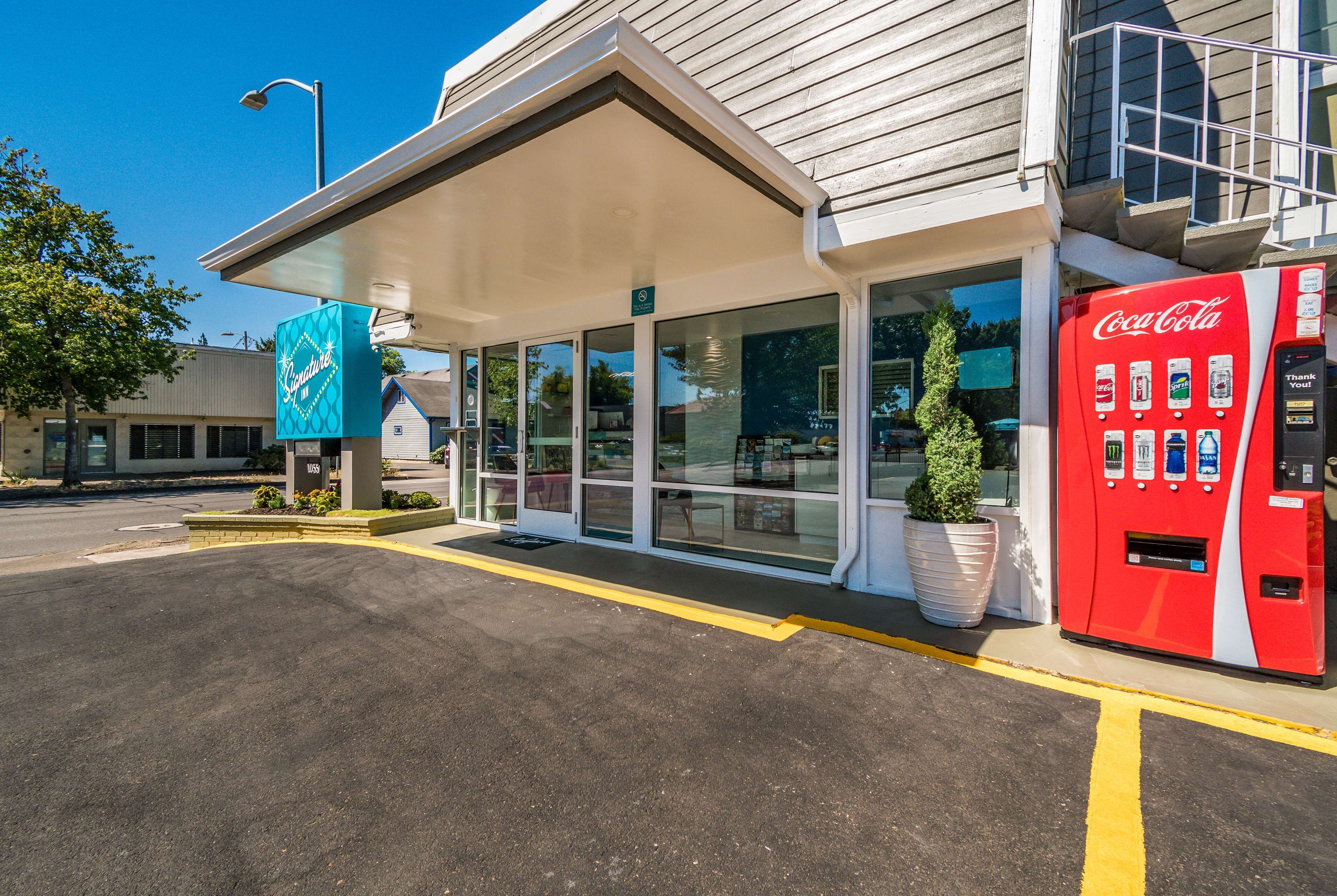 A building with a Coca-Cola vending machine, a covered entrance, and a large potted plant.