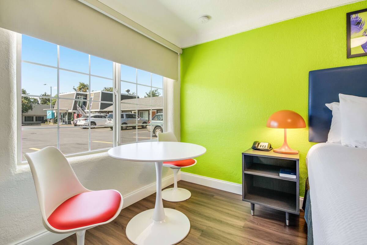 A hotel room with a bed, a small table, two chairs, a nightstand, and a window with a view of a parking lot and buildings.