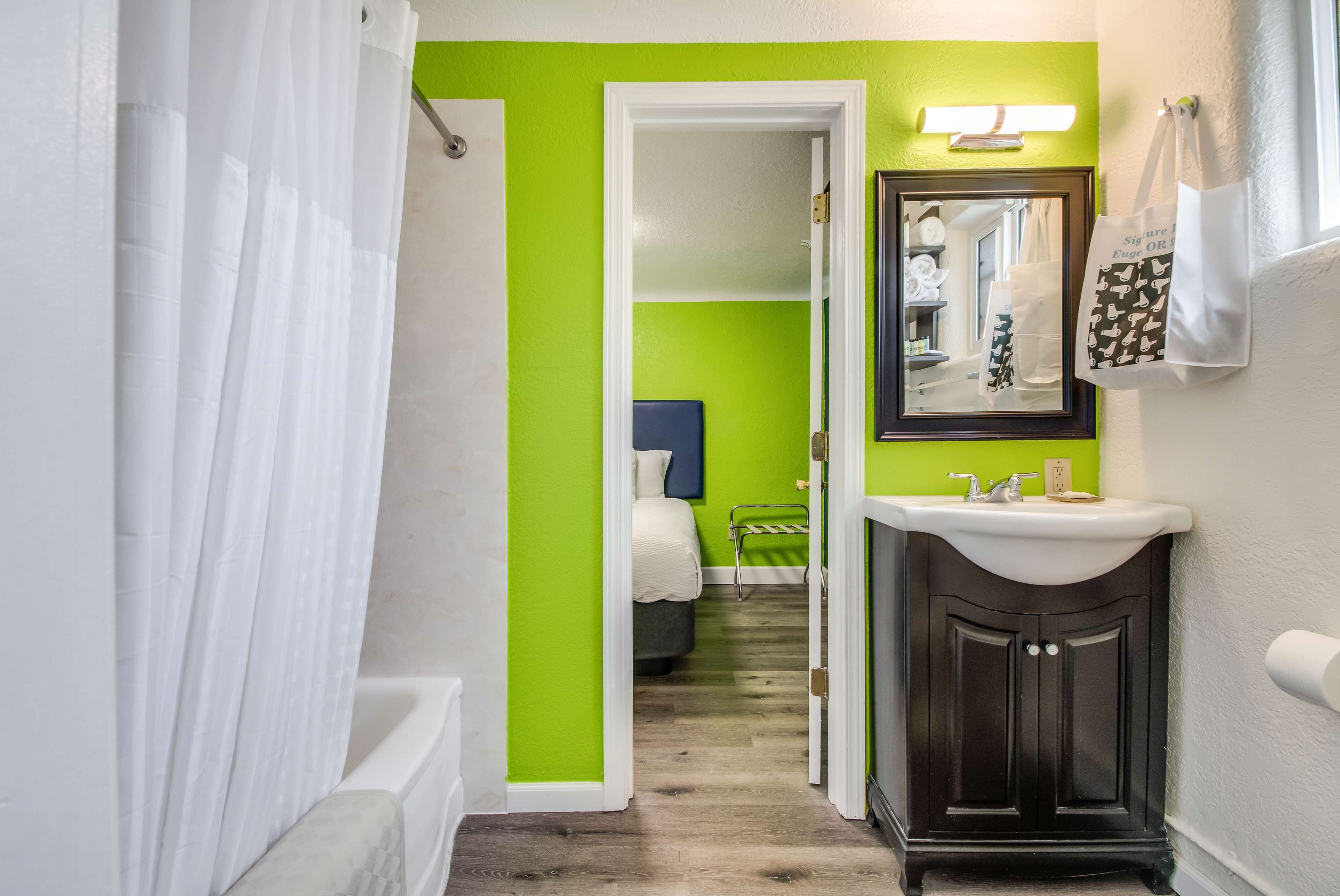 Une salle de bain avec un mur d’accent vert, une baignoire blanche, un meuble-lavabo en bois foncé avec un miroir, et une serviette blanche accrochée au mur.