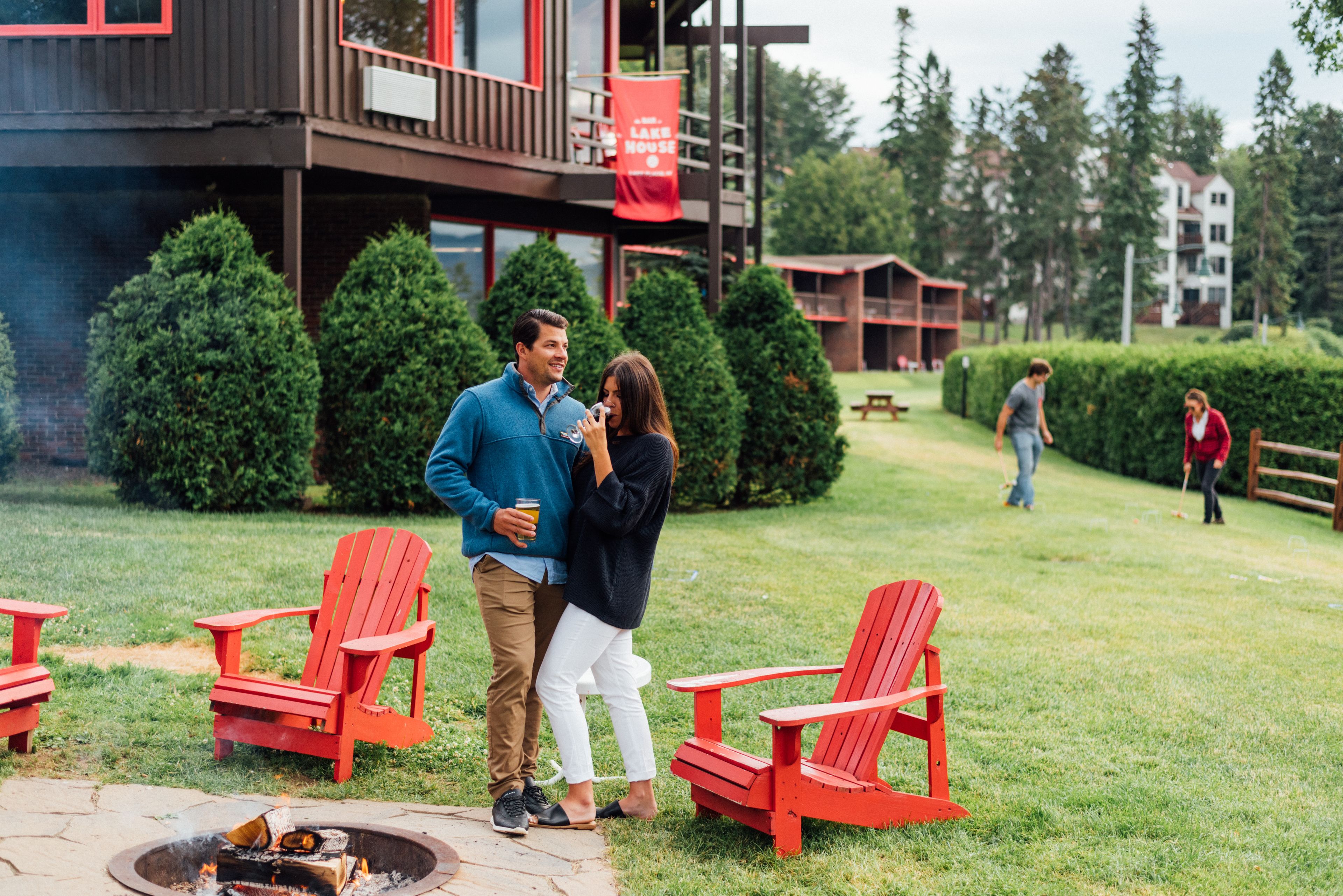 A couple standing by a fire pit with red Adirondack chairs, a building with a red sign, and a grassy area with trees and a wooden fence in the background.
