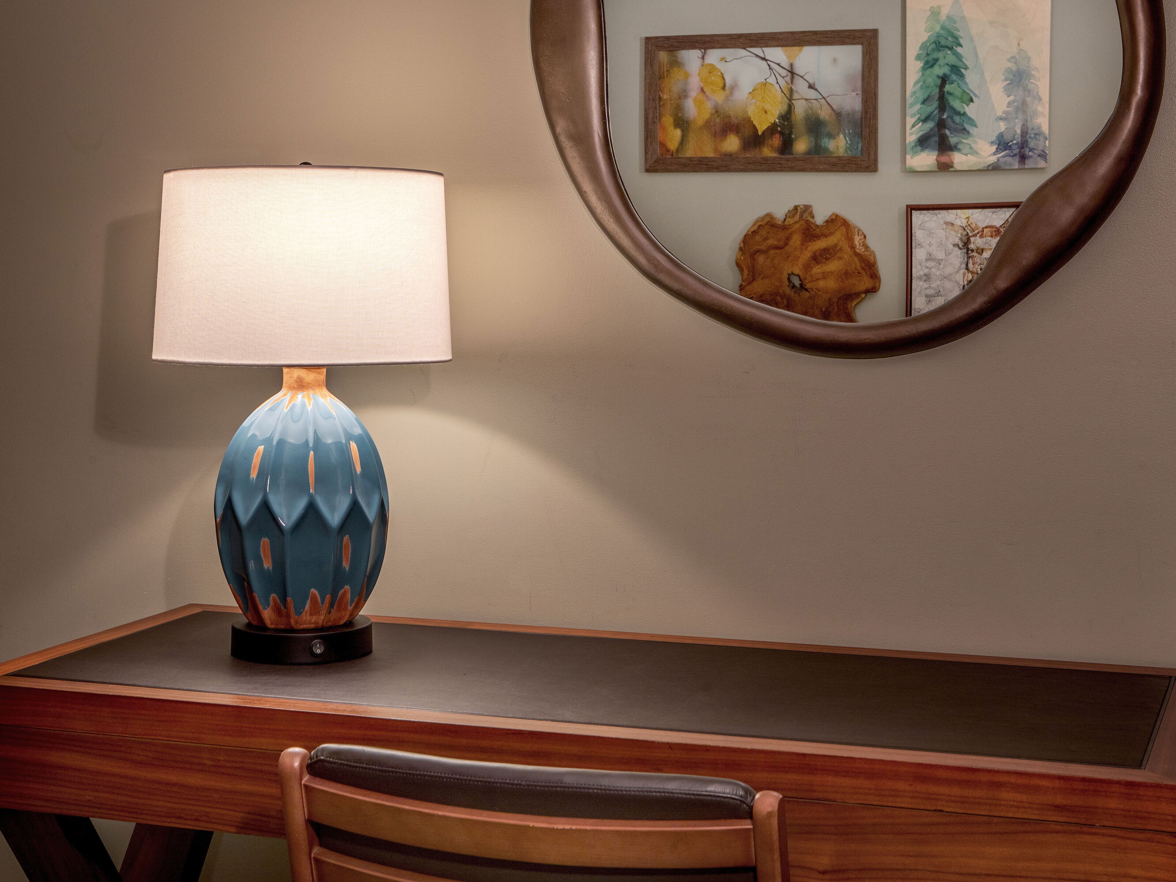 A desk with a lamp, a chair, and a mirror reflecting artwork.