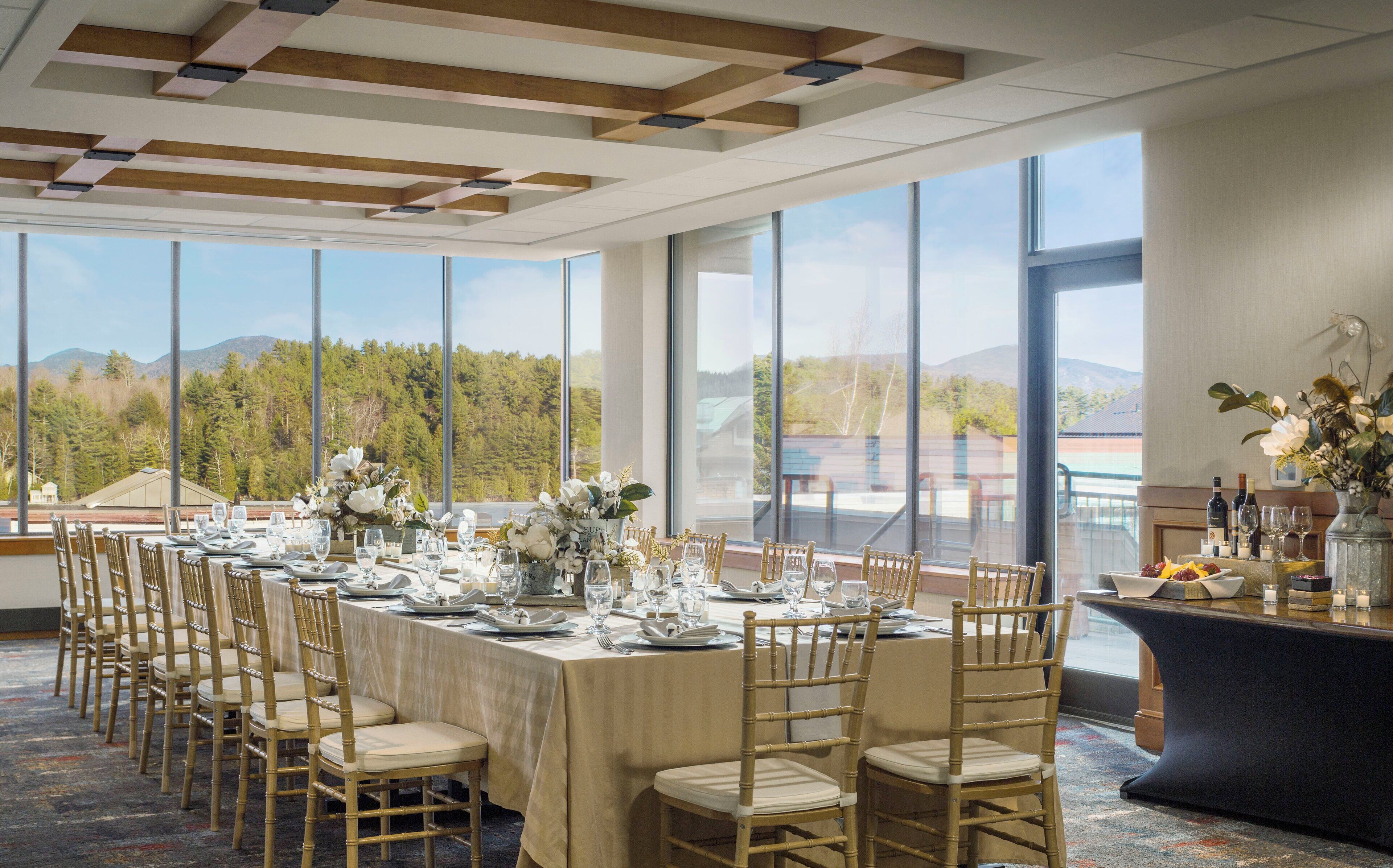 A banquet hall with a long table set for a formal event, large windows offering a scenic mountain view, and a modern ceiling design.