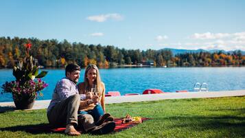 Un couple est assis sur une couverture près d’un lac, avec une piscine et des chaises longues en arrière-plan.