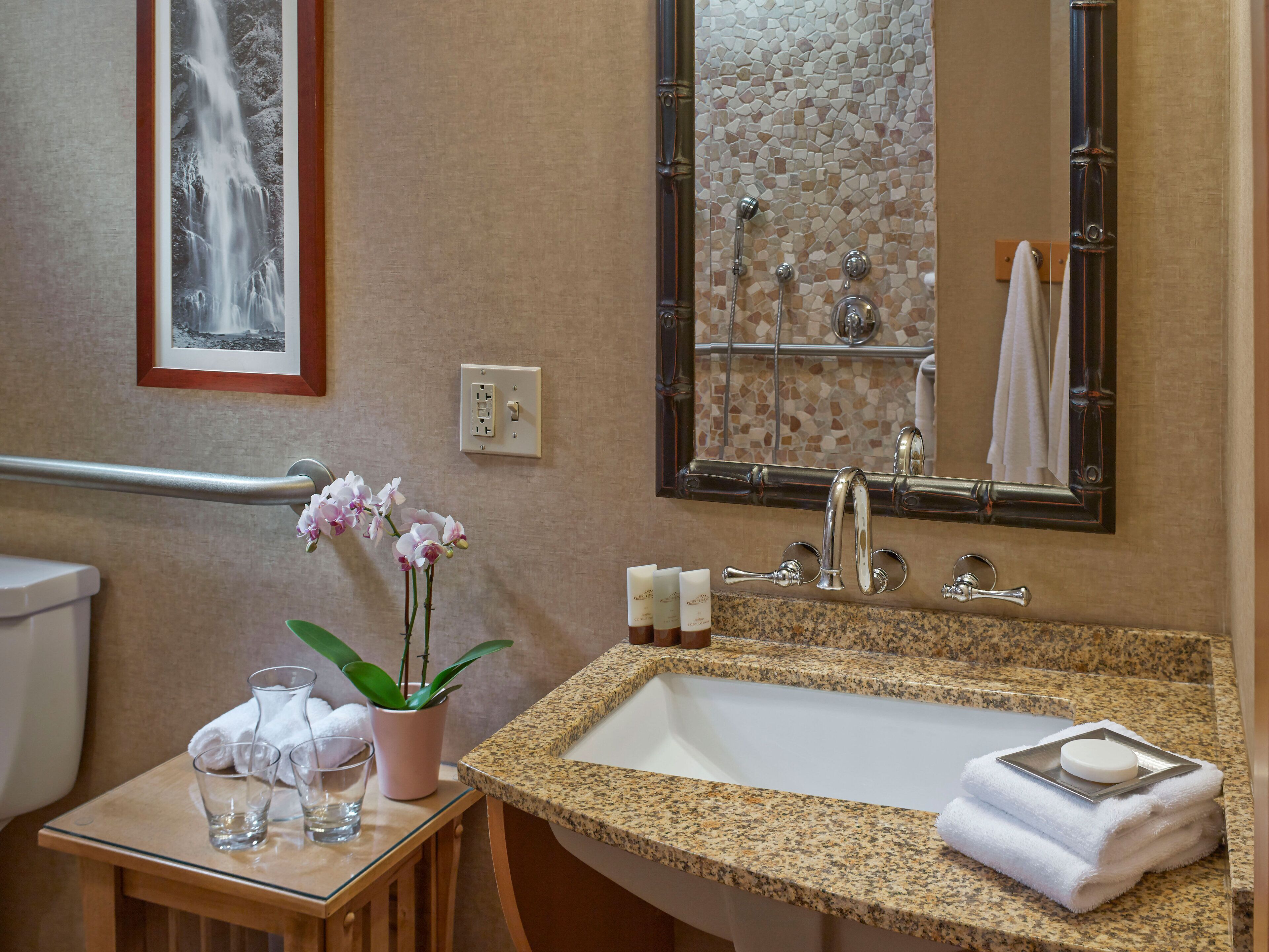 A bathroom with a granite sink, a mirror, a towel rack, a framed picture, and a small table with a plant and glasses.