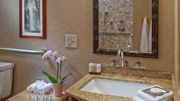 A bathroom with a granite sink, a mirror, a towel rack, a framed picture, and a small table with a plant and glasses.
