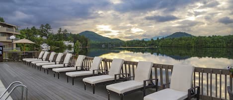 A deck with lounge chairs facing a lake, surrounded by trees and hills.