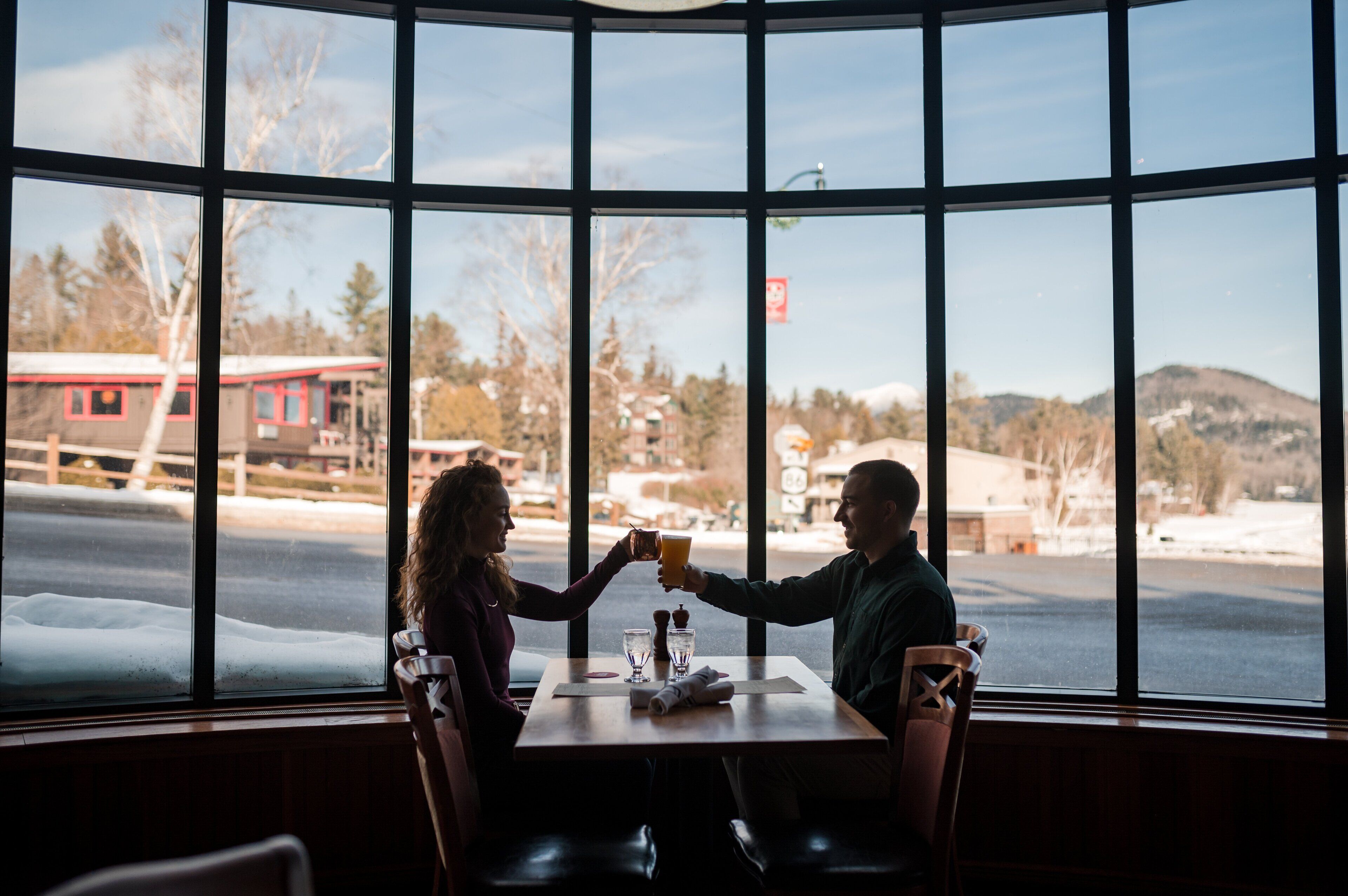 Un couple trinque à une table dans un restaurant aux grandes fenêtres donnant sur un paysage enneigé.
