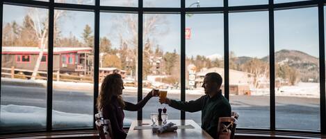 Un couple trinque à une table dans un restaurant aux grandes fenêtres donnant sur un paysage enneigé.