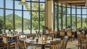 A restaurant with large windows offering a view of a lake and mountains. Tables are set with chairs and dining ware. - High Peaks Resort (Lake Placid)