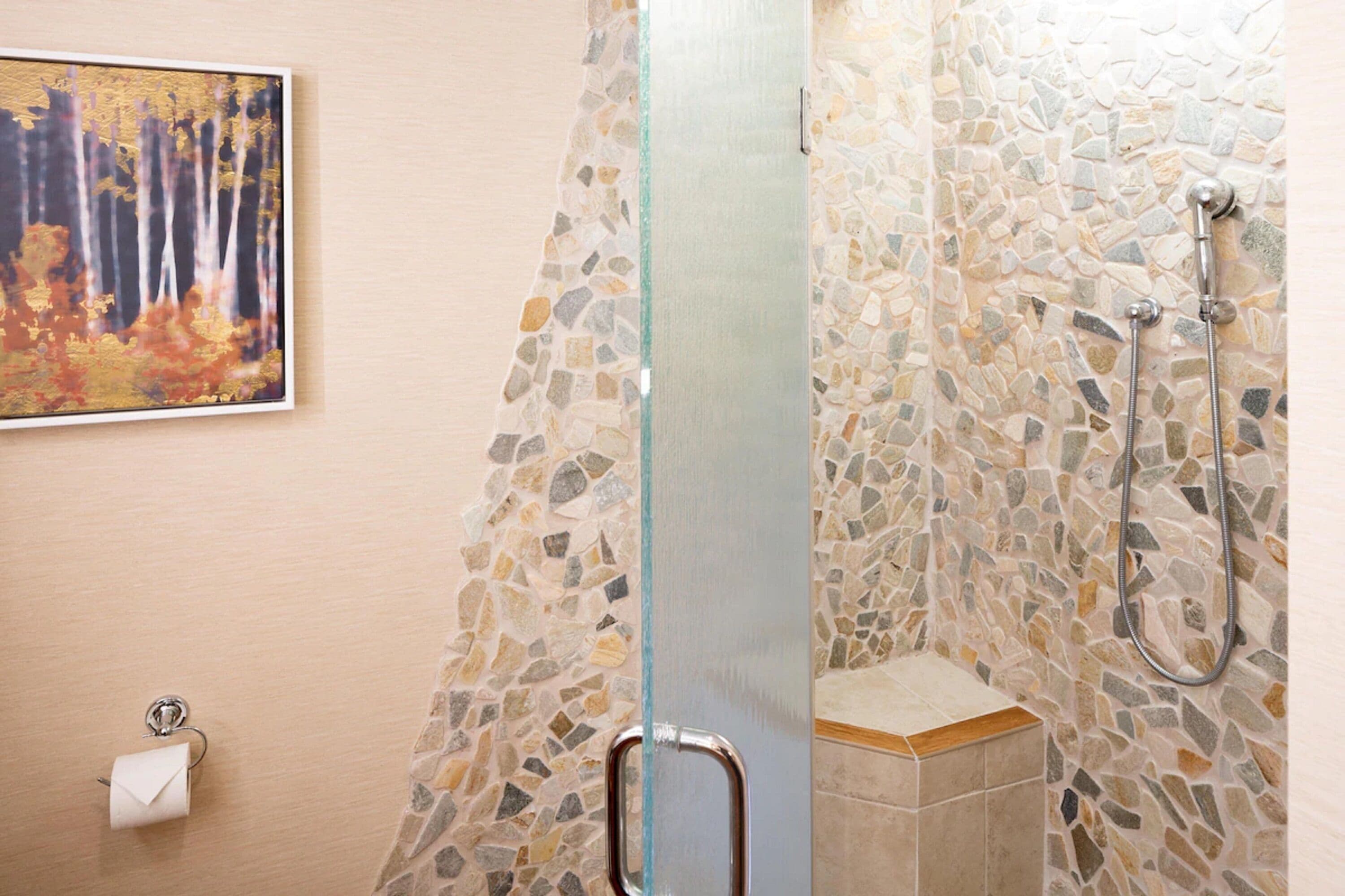 A bathroom with mosaic tile walls, a glass shower door, a toilet paper holder, and a showerhead.