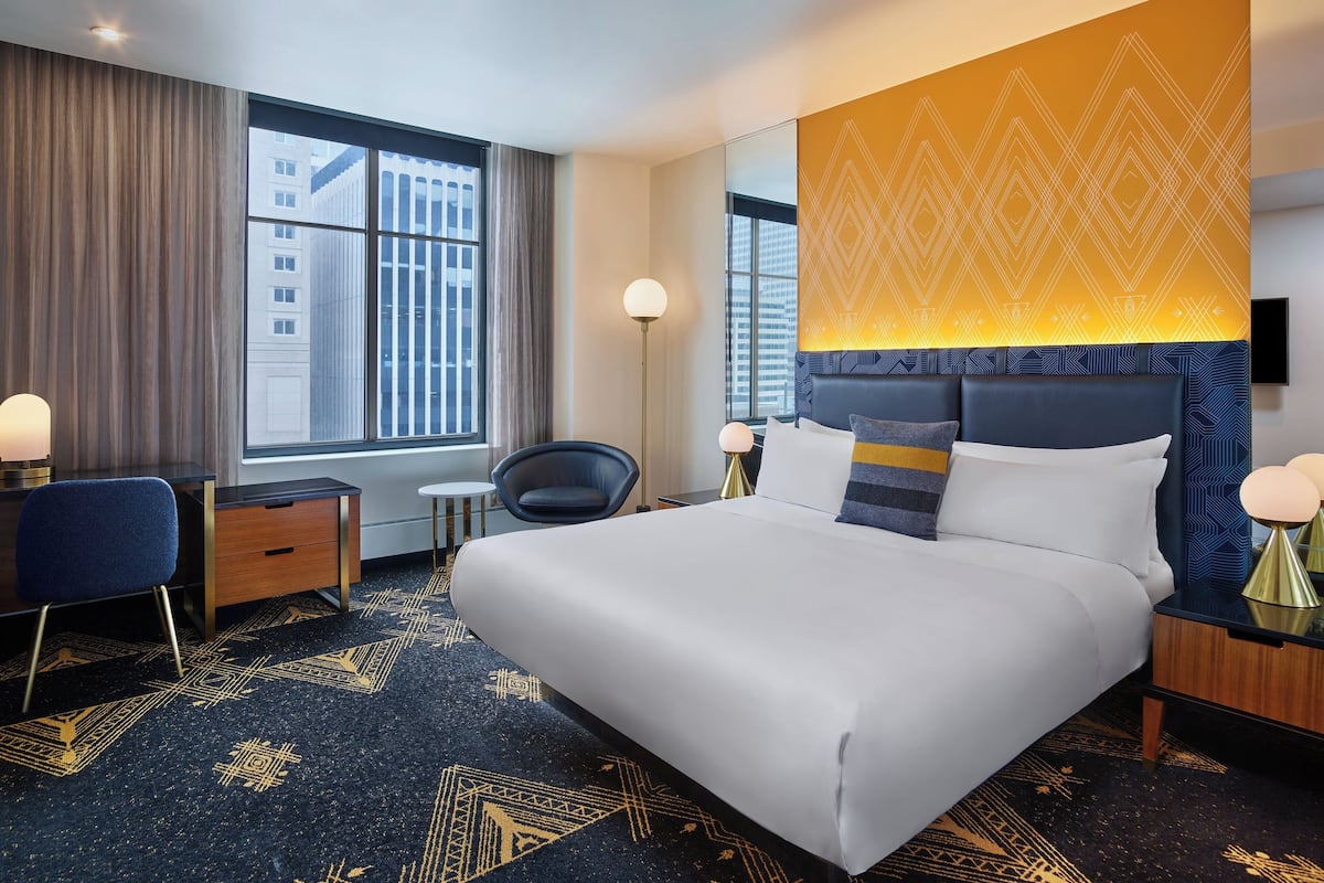 Quarto de hotel com uma cama grande, uma escrivaninha, uma cadeira e uma janela ampla com vista para edifícios.