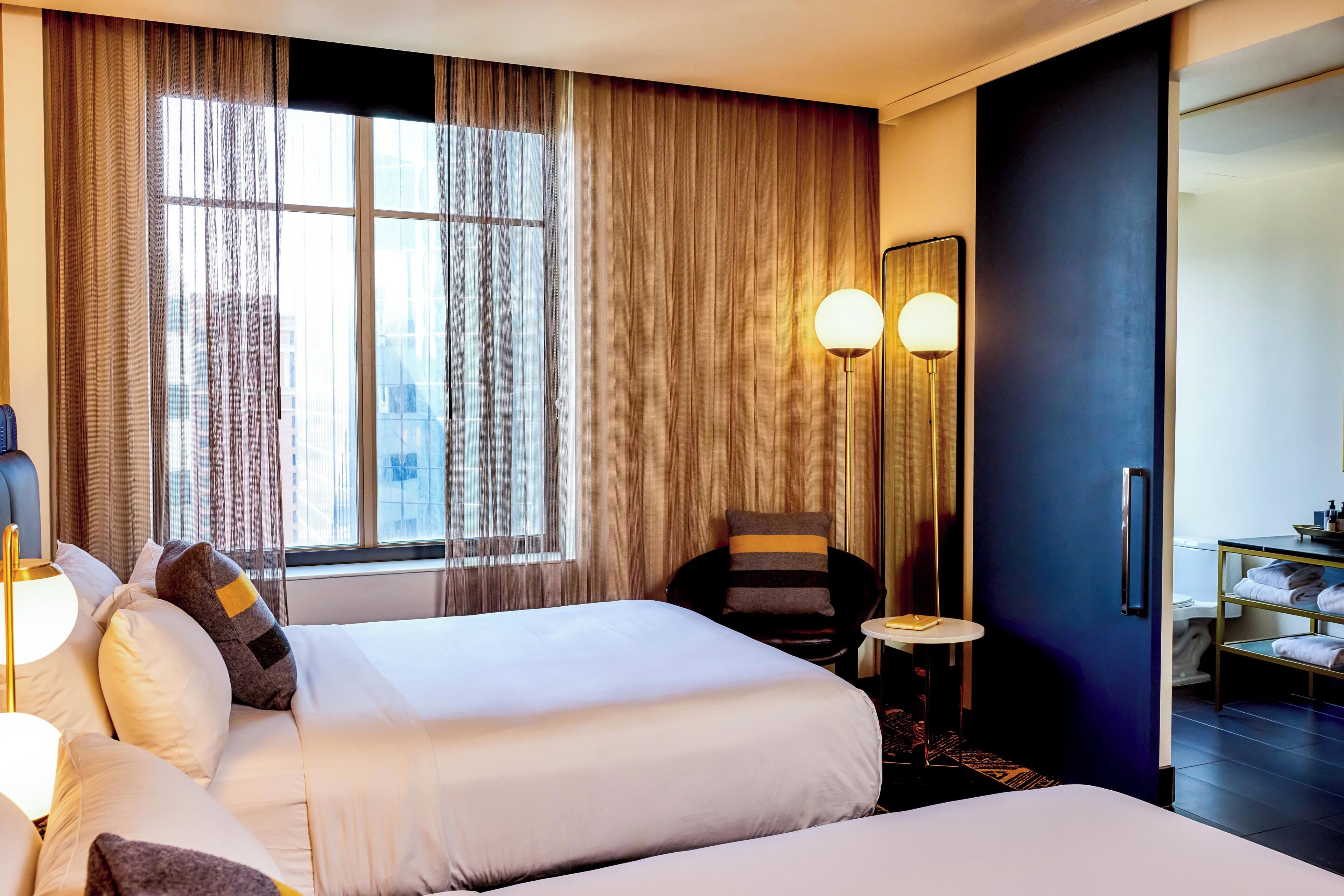 Une chambre d’hôtel avec deux lits, un coin salon comprenant un fauteuil, une petite table et une grande fenêtre avec des rideaux.