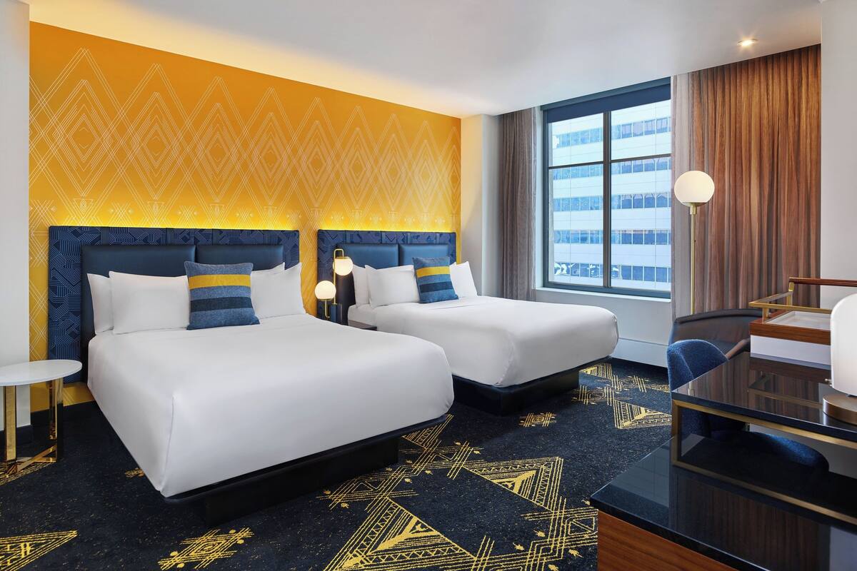 Quarto de hotel com duas camas, uma escrivaninha e uma janela grande.