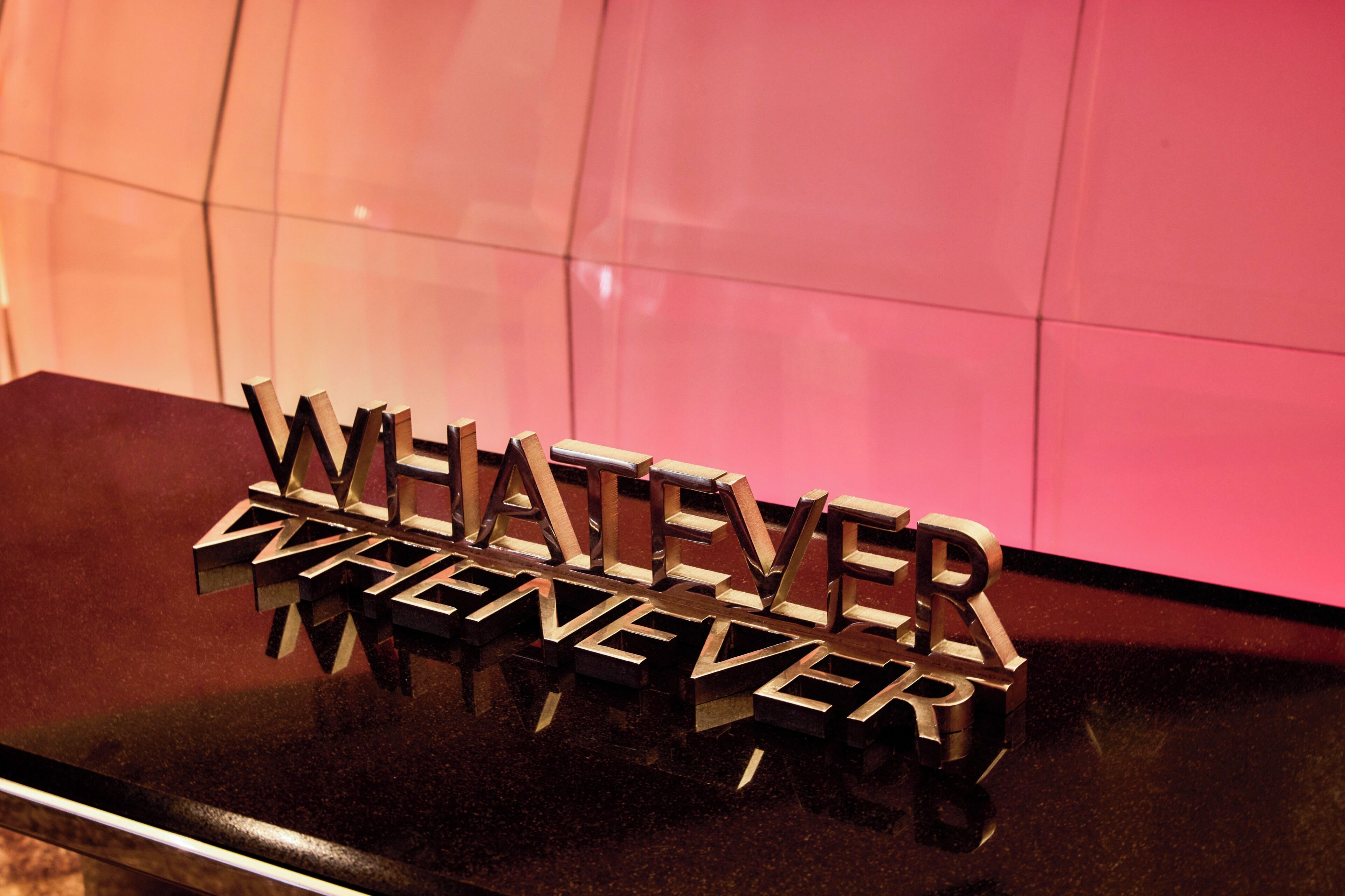 Ein metallenes Schild mit der Aufschrift 'WHATEVER WHENEVER' auf einer spiegelnden Oberfläche.