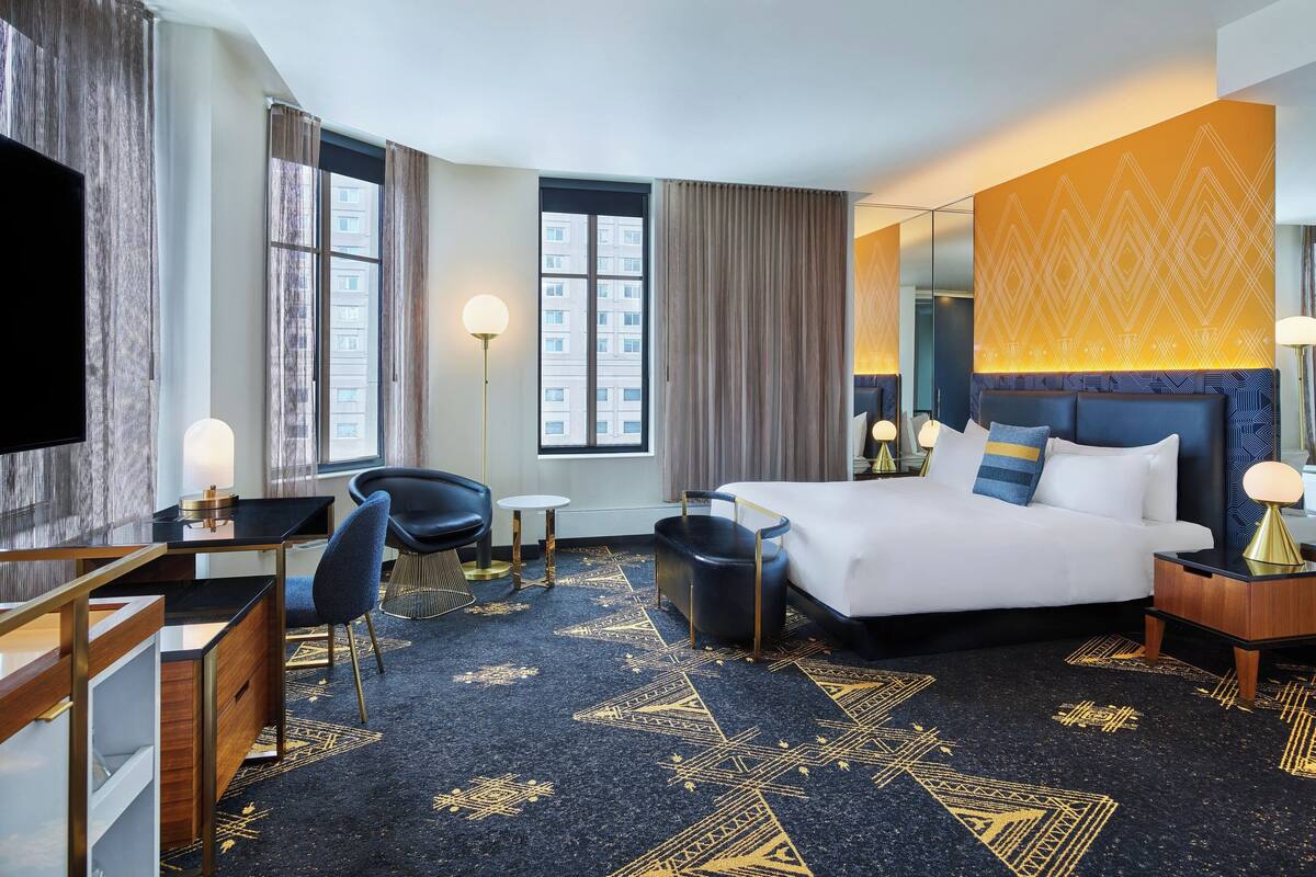Quarto de hotel com cama, escrivaninha, cadeira e uma janela grande.