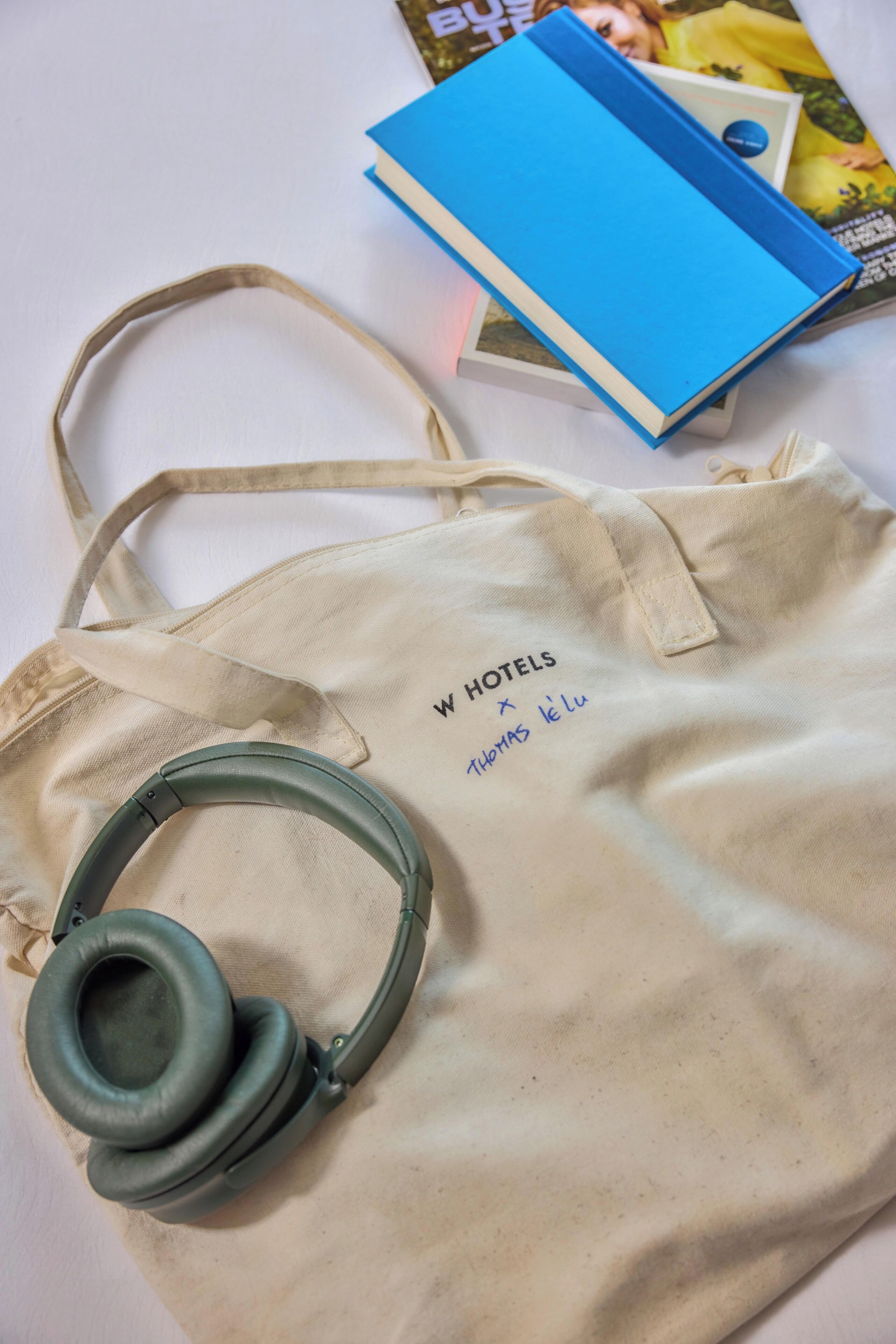Una borsa porta tutto beige con la scritta 'W HOTELS' e 'THOMAS KELU', una cuffia e un libro blu con una copertina di rivista.
