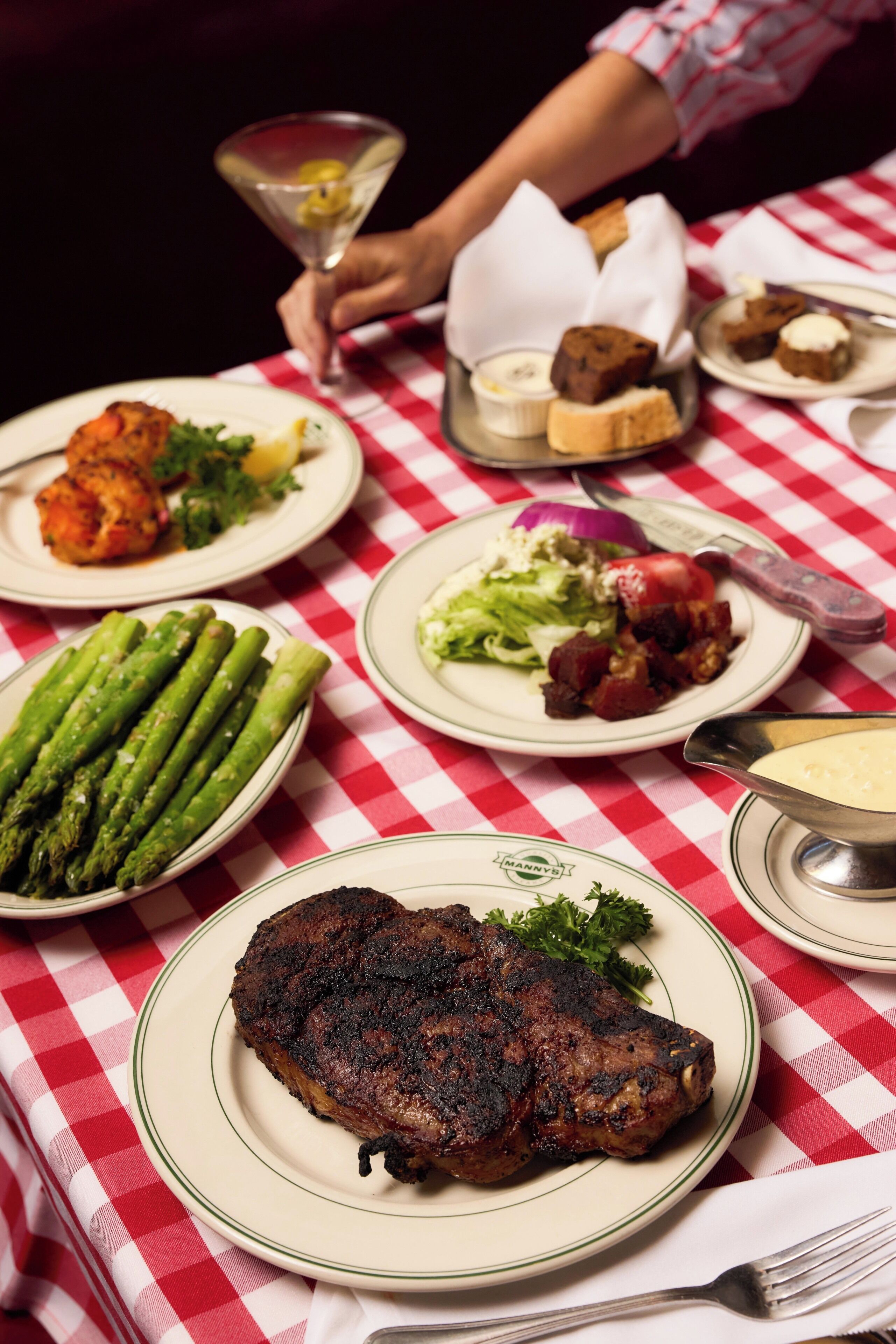Una mesa con varios platos, incluyendo espárragos, un bistec y una copa de martini.