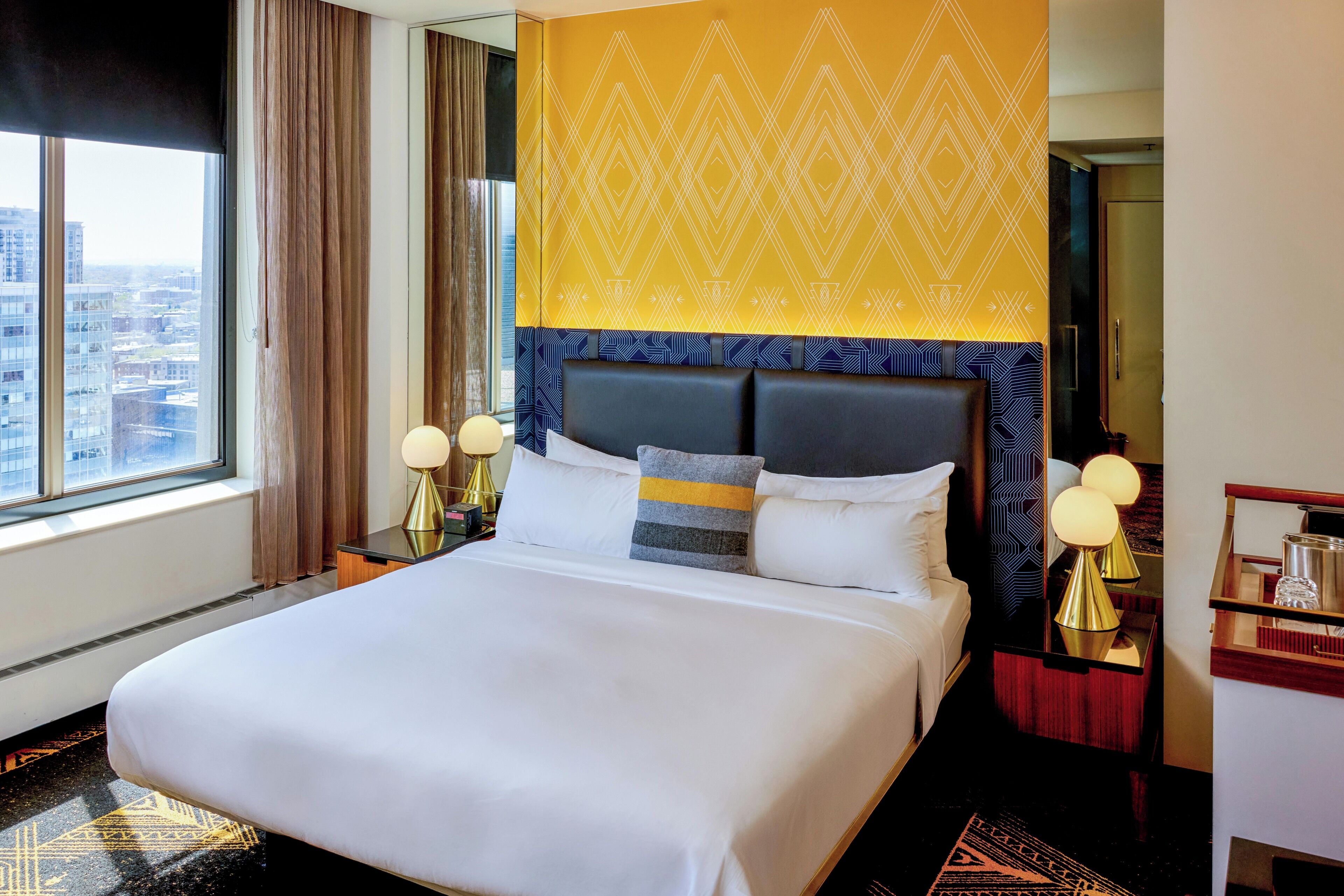 A hotel room with a large bed, a city view, a patterned headboard, and a nightstand with lamps.