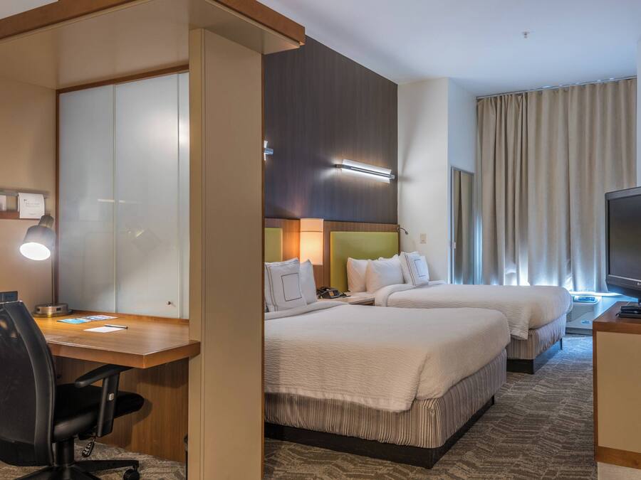 A hotel room with two beds, a desk with a chair, a television, and a large window with curtains.