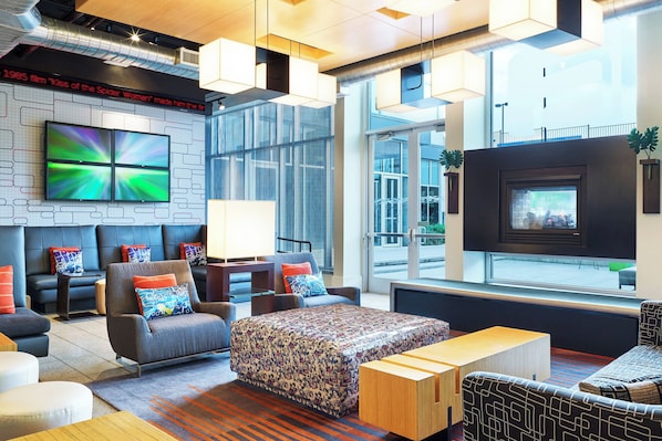 A modern hotel lobby with a seating area, a flat-screen TV, a fireplace, and large windows. - Aloft by Marriott Las Colinas (Irving)