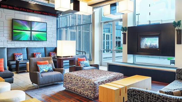 A modern lounge area with comfortable seating, a large flat-screen TV, and a fireplace. - Aloft by Marriott Las Colinas (Irving)