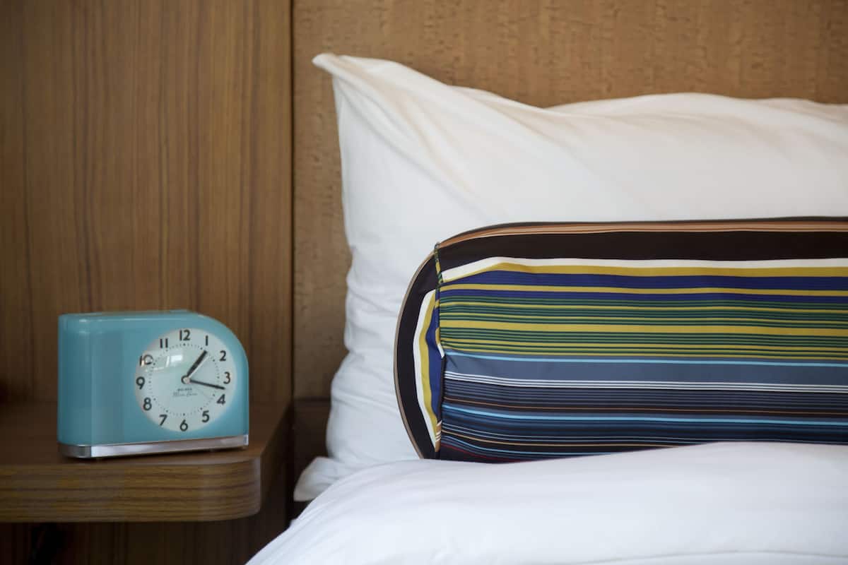 Una cama con sábanas blancas y un cojín a rayas, un reloj azul sobre una mesita de noche de madera, y un cabecero de madera.