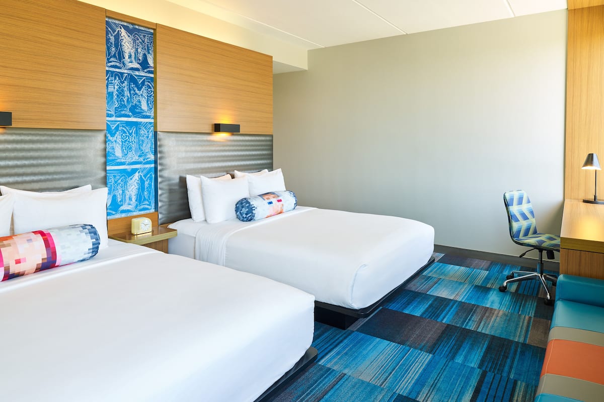 A hotel room with two beds, a desk, a chair, and a large wall tapestry.