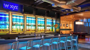 Zona de bar con un letrero de neón, varias estanterías llenas de botellas y taburetes.
