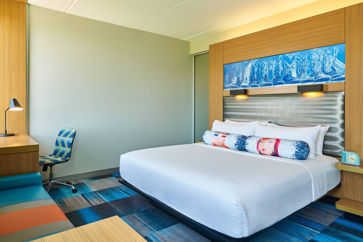 A hotel room with a large bed, a desk with a chair, a wall-mounted artwork, and a patterned carpet.