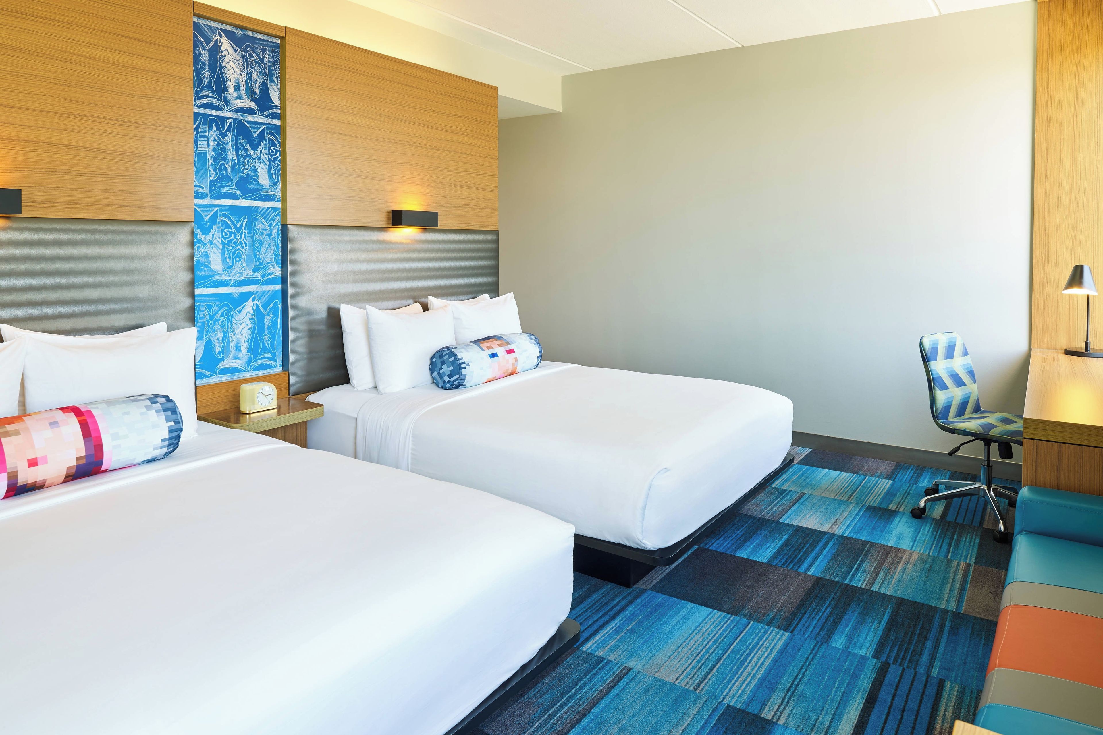 A hotel room with two beds, a desk, a chair, and a patterned carpet.