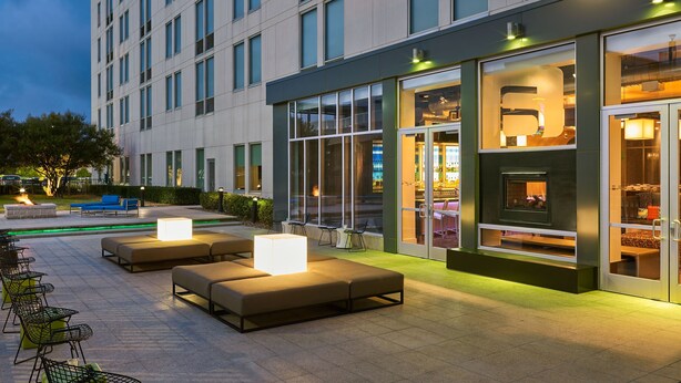 Un espacio de estar al aire libre con muebles modernos y una chimenea, junto a un edificio con ventanas amplias y un puerta de vidrio.