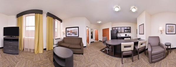 Quarto de hotel com televisão, sofá, poltrona, mesa de jantar e área de cozinha.