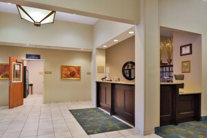A hotel lobby with a reception desk, a coffee machine, a trash can, a framed artwork, and a carpeted floor.