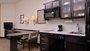 A compact hotel room with a kitchenette, including a microwave, coffee maker, and sink. There is a desk with a chair and a flat-screen TV.